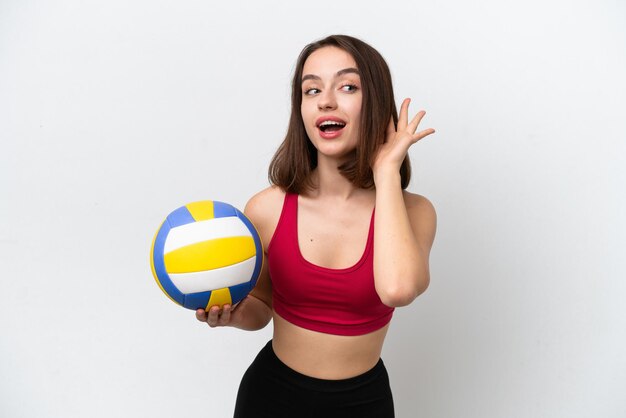 Junge ukrainische Frau spielt Volleyball isoliert auf weißem Hintergrund und hört etwas, indem sie die Hand auf das Ohr legt