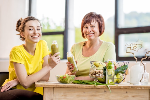 Junge und ältere Frauen, die nach dem Sporttraining drinnen auf dem Fensterhintergrund mit gesundem Essen und frischen Getränken sitzen