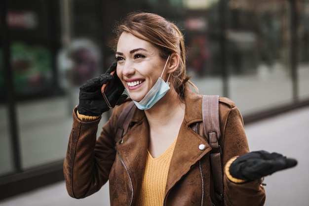 Junge und elegante Geschäftsfrau, die auf leere Stadtstraße geht. Sie benutzt das Telefon, während sie eine Schutzmaske trägt, um sich vor gefährlichen Viren zu schützen. Corona-Virus oder Covid-19-Konzept.