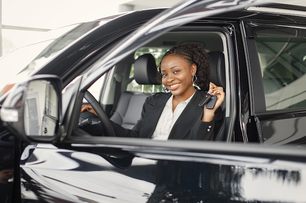 Junge und fröhliche Frau, die neues Auto genießt, während sie im Inneren sitzt Schwarze Frau, die einen Autoschlüssel hält Mädchen, das schwarzes Kostüm trägt