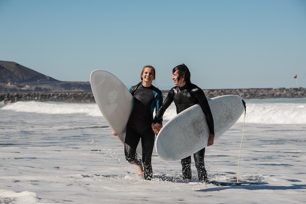 Junge und glücklich lächelnde paar Surfer in schwarzen Neoprenanzügen, die sich an den Händen halten und mit Surfbrettern im Wasser gehen