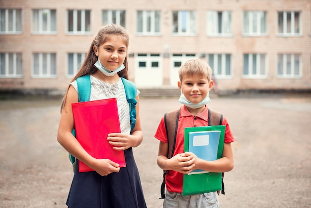 Junge und Mädchen mit Schultaschen, die medizinische Masken unter dem Kinn tragen und Lernmaterialien halten