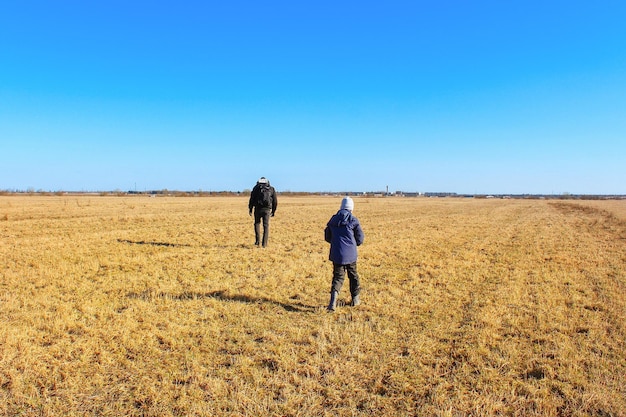 Junge und Mann gehen auf trockenem Grasfeld unter blauem Himmel, Blick von hinten, sonniger Tag