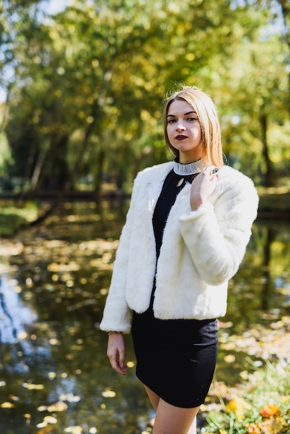 Junge und schlanke Frau mit schwarzem Kleid und Pelzjacke posiert im Herbstpark