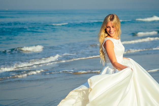 Junge und schöne Braut mit dem langen blonden Haar, das auf dem Ozeanhintergrund in einem schönen weißen langen Hochzeitskleid lächelt und geht. eine glückliche Braut heiratete eine Hochzeitszeremonie am Strand am Meer