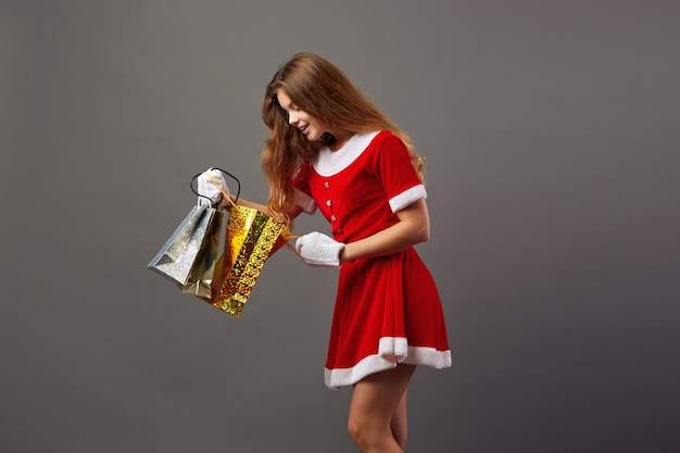 Junge und schöne Frau Santa Claus mit Sonnenbrille in der roten Robe und weißen Handschuhen hält die Taschen mit Geschenken und schaut in sie auf dem grauen Hintergrund.