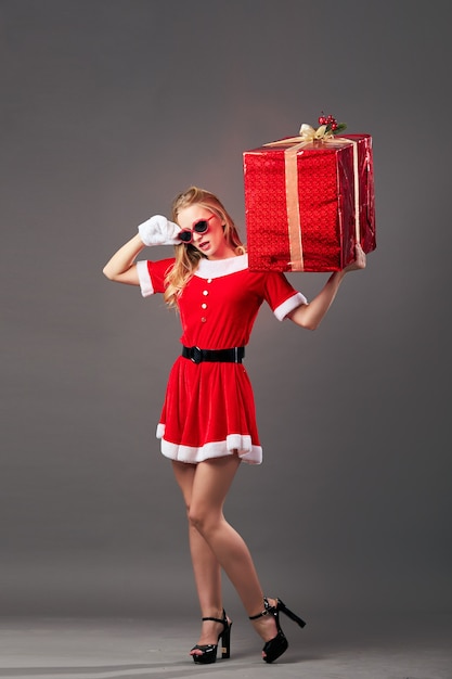 Junge und schöne Frau Santa Claus mit Sonnenbrille in der roten Robe, weißen Handschuhen und High Heels hält das riesige Weihnachtsgeschenk auf dem grauen Hintergrund. .