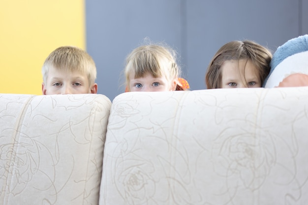 Junge und zwei Mädchen verstecken sich hinter dem Sofa und schauen hinaus.