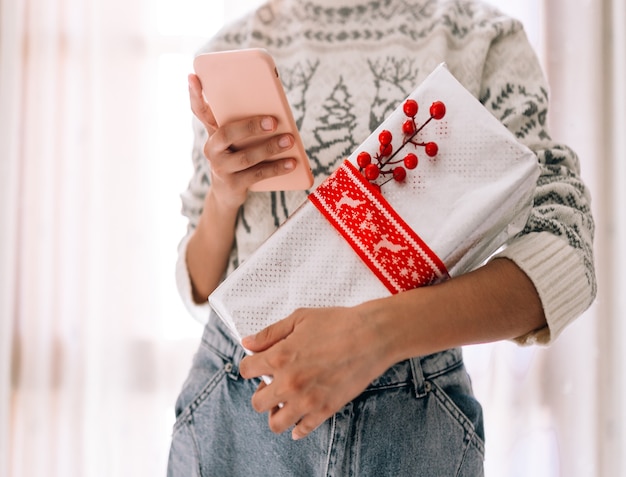 Foto junge unerkennbare frau, die ihr handy beobachtet, hält in einer geschenkbox in metallischem papier