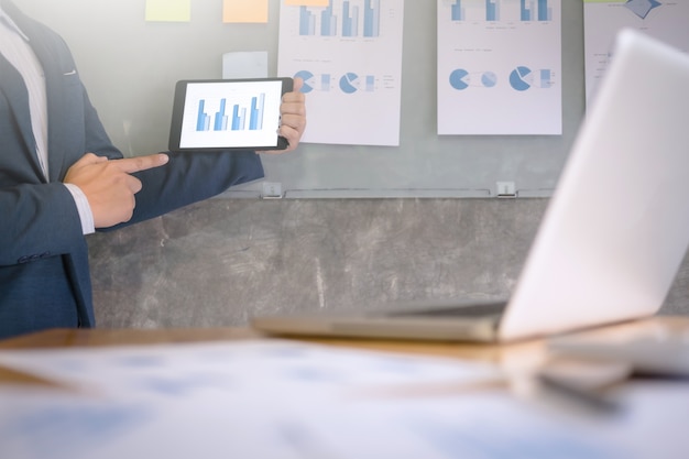 Junge Unternehmer in der Anzug erklären Daten Diagramm auf digitale Tablette und Dokumente an der Wand in einem Büro.