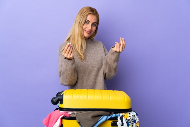 Junge uruguayische blonde Frau mit einem Koffer voller Kleidung über isolierter lila Wand, die Geldgeste macht