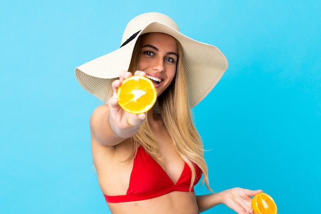 Junge uruguayische blonde Frau über isoliertem blauem Hintergrund im Badeanzug und hält eine Orange