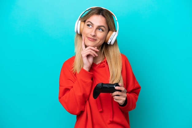 Junge uruguayische Frau, die mit einem Videospiel-Controller spielt, isoliert auf Blau, denkt eine Idee, während sie nach oben schaut