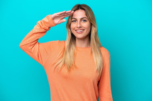 Junge uruguayische Frau isoliert auf blauem Hintergrund, die mit der Hand mit glücklichem Ausdruck salutiert