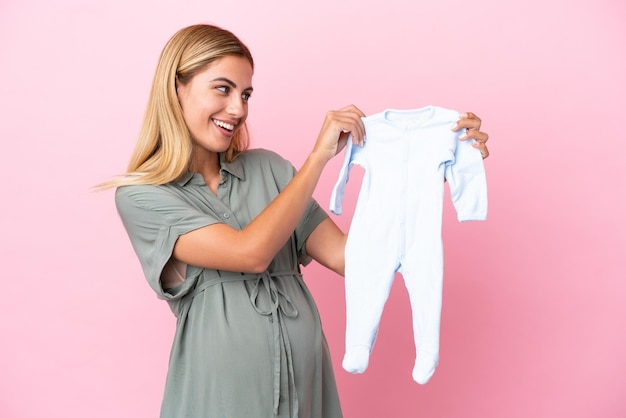 Junge uruguayische Frau isoliert auf blauem Hintergrund schwanger und hält Babykleidung