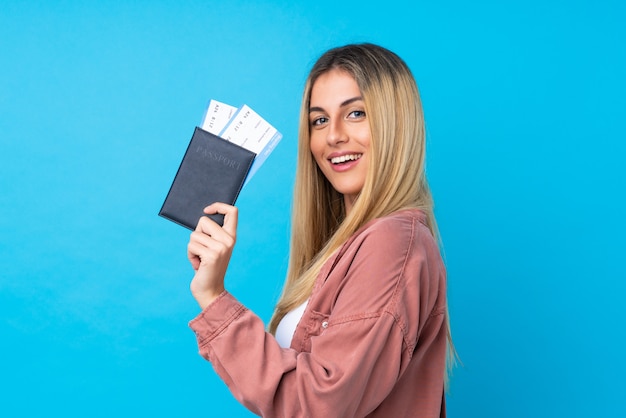 Junge uruguayische Frau über isolierte blaue Wand glücklich im Urlaub mit Pass und Flugtickets