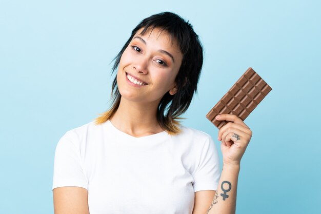 Junge uruguayische Frau über isoliertem Blau, das eine Schokoladentafel nimmt und glücklich ist