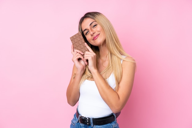 Junge uruguayische Frau über isolierter rosa Wand, die eine Schokoladentafel nimmt und glücklich ist