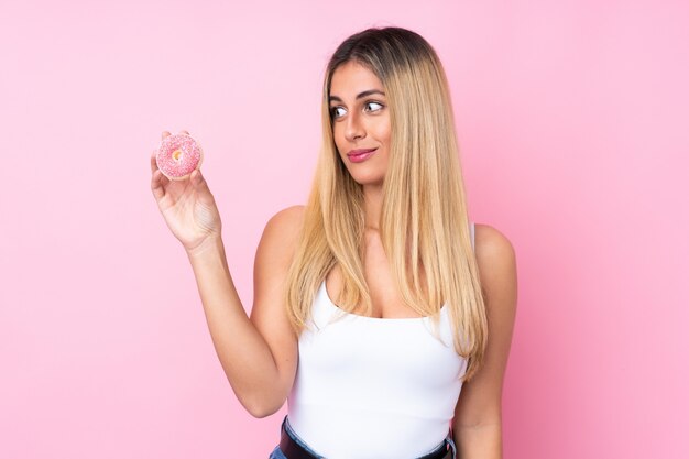 Junge uruguayische Frau über isolierter rosa Wand, die einen Donut hält