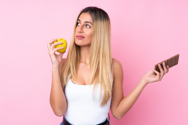 Junge uruguayische Frau über isolierter Wand rosa Wand, die Zweifel hat, während sie eine Schokoladentafel in einer Hand und einen Apfel in der anderen nimmt