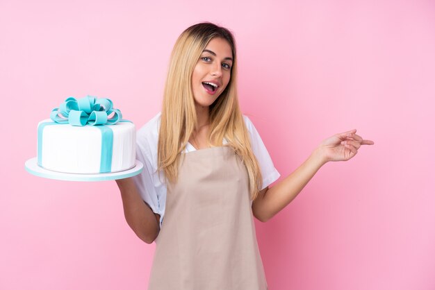 Junge uruguayische Gebäckfrau mit einem großen Kuchen über isolierter Wand rosa Wand überrascht und Finger zur Seite zeigend