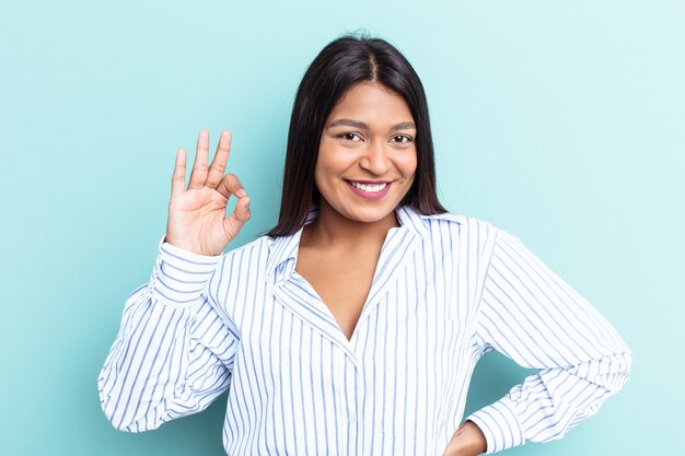 Junge venezolanische Frau, die auf blauem Hintergrund isoliert ist, zwinkert ein Auge und hält eine okaygeste mit der Hand.