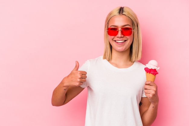 Junge venezolanische Frau, die ein Eis isst, das auf rosa Wand lokalisiert wird, die lächelt und Daumen aufhebt