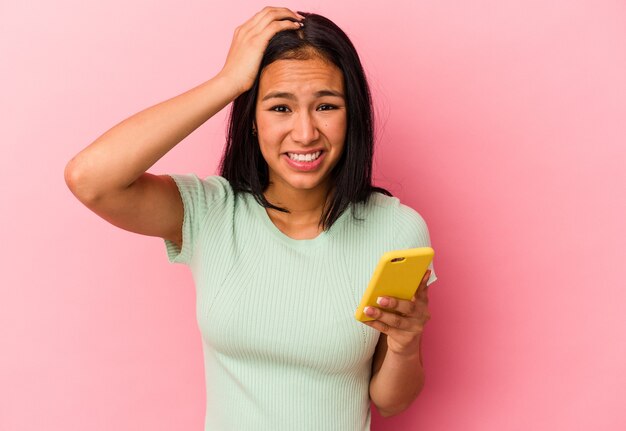 Junge venezolanische Frau, die ein Mobiltelefon einzeln auf rosafarbenem Hintergrund hält und schockiert ist, hat sich an ein wichtiges Treffen erinnert.