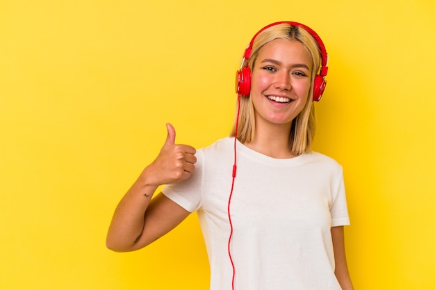 Junge venezolanische Frau, die Musik hört, die auf gelbem Hintergrund lokalisiert wird, der lächelt und Daumen aufhebt