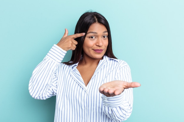 Junge venezolanische Frau isoliert auf blauem Hintergrund, die ein Produkt zur Hand hält und zeigt.