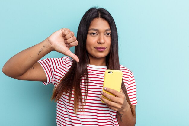 Junge venezolanische Frau mit Handy isoliert auf blauem Hintergrund mit einer Abneigungsgeste, Daumen nach unten. Meinungsverschiedenheit Konzept.