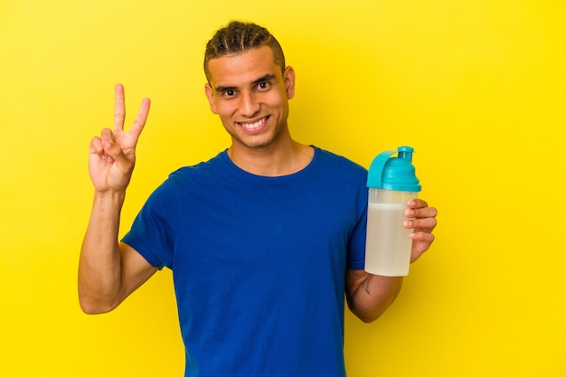 Junge venezolanische Mann, der einen Proteinshake trinkt, der auf gelbem Hintergrund isoliert ist und Nummer zwei mit den Fingern zeigt.