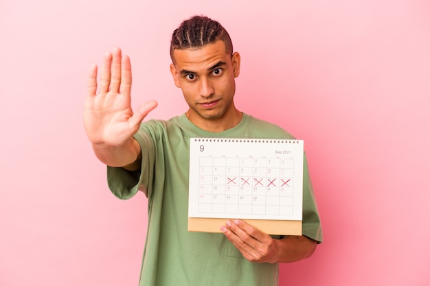 Junge venezolanische Mann hält einen Kalender isoliert auf rosa Hintergrund stehend mit ausgestreckter Hand mit Stoppschild und verhindert Sie.