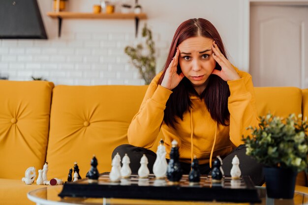 Junge verärgerte Frau, die Schach spielt und auf dem Sofa sitzt Distressed weibliche Spiele im logischen Brettspiel mit sich selbst im Zimmer