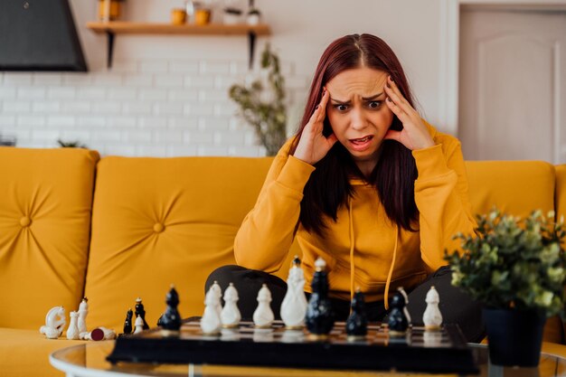 Junge verärgerte Frau, die Schach spielt und auf dem Sofa sitzt Distressed weibliche Spiele im logischen Brettspiel mit sich selbst im Zimmer