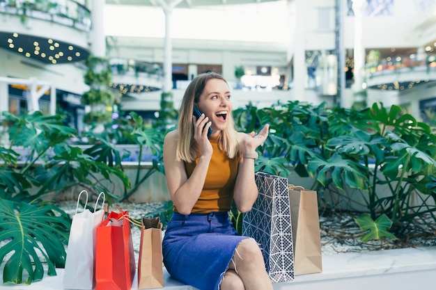 Junge Verbraucherin im Einkaufszentrum durchsucht den Chat und verwendet ein Smartphone. Frau, die mit einem Handy in den Händen im Einkaufszentrum steht. drinnen. Happy Shopper Girl mit Geschenktüten macht Einkäufe