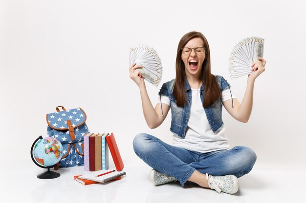 Junge verrückte Studentin, die schreit, Hände halten Bündel viele Dollar, Bargeld sitzt in der Nähe von Globus Rucksack, Bücher isoliert