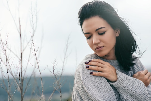 Junge verträumte Frau mit geschlossenen Augen mit der Hand auf der Schulter, die einen grauen Pullover trägt Attraktive brünette kaukasische junge Frau mit windigem Haar im Freien Lifestyle-Modekonzept Cover-Idee