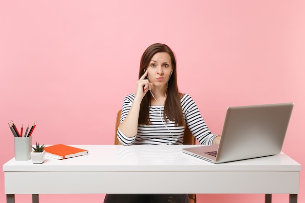 Junge verwirrte Frau mit Kopfhörern, die Musik Hörbuch hört, sitzt und arbeitet am weißen Schreibtisch mit zeitgenössischem PC-Laptop einzeln auf pastellrosa Hintergrund. Erfolgsgeschäftskarrierekonzept. Platz kopieren.