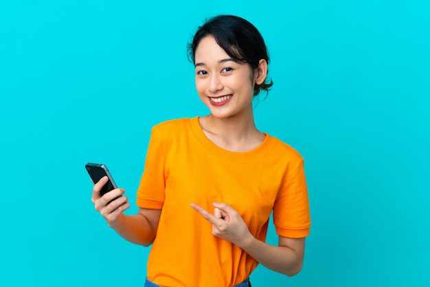 Junge vietnamesische Frau isoliert auf blauem Hintergrund mit Handy und zeigt darauf