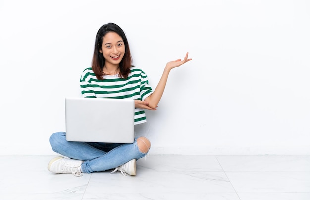 Junge vietnamesische Frau mit einem Laptop, der auf dem Boden sitzt, isoliert auf einer weißen Wand, die die Hände zur Seite ausstreckt, um zum Kommen einzuladen