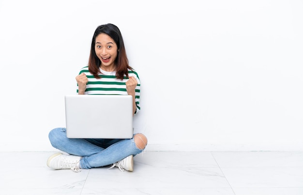 Junge vietnamesische Frau mit einem Laptop, der isoliert auf einer weißen Wand auf dem Boden sitzt und einen Sieg in Siegerposition feiert