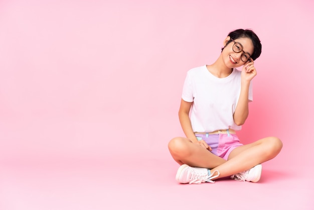 Junge vietnamesische Frau mit kurzen Haaren, die auf dem Boden über isolierter rosa Wand mit Brille und glücklich sitzen