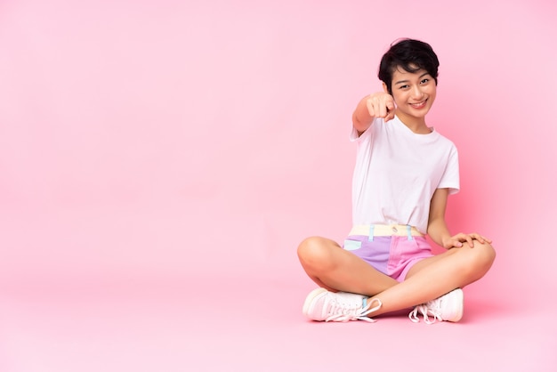 Junge vietnamesische Frau mit kurzen Haaren, die auf dem Boden über isolierter rosa Wand sitzen, zeigt Finger auf Sie mit einem selbstbewussten Ausdruck