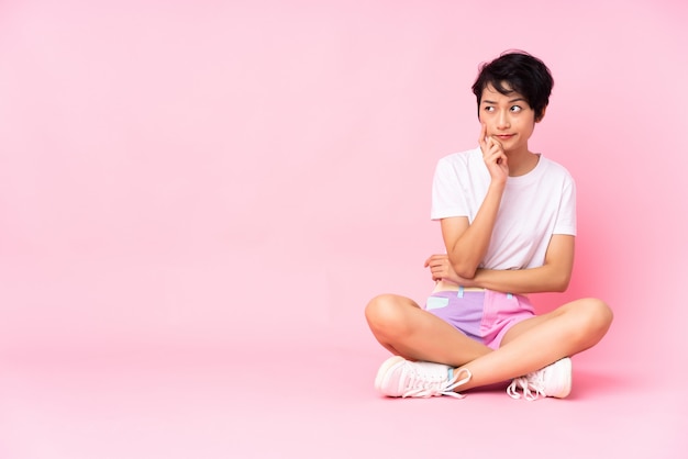 Junge vietnamesische Frau mit kurzen Haaren, die auf dem Boden über rosa Wand sitzen, die Zweifel und mit verwirrendem Gesichtsausdruck hat