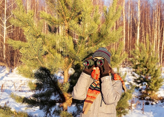 Junge Vogelbeobachterin in Winterkleidung und Strickschal, die durch ein Fernglas im Wald schaut