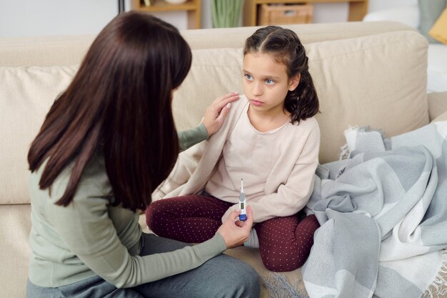 Junge vorsichtige Frau, die sich um ihre kranke, entzückende Tochter kümmert, die auf der Couch sitzt, während sie ihre Schulter berührt, nachdem sie die Körpertemperatur gemessen hat