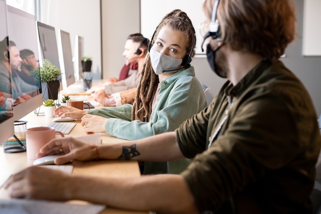 Junge weibliche Betreiberin des Callcenters in Schutzmaske, die einen der Kollegen anschaut, während sie sich mit ihm über Kundenfragen berät