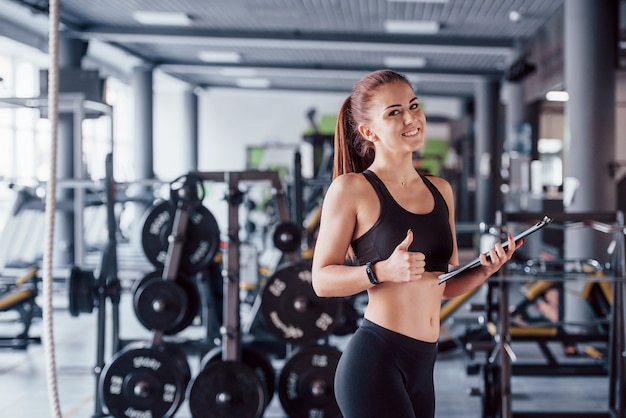 Junge weibliche Fitness Personal Trainer mit Notizblock in der Turnhalle mit Daumen nach oben stehen.