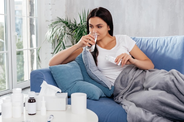 Junge weibliche Trinkwasser nach der Einnahme von Pillen, während sie sich auf der Couch ausruhen